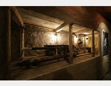 Ein rustikaler, dimm beleuchteter Raum mit Lehmwänden und Holzbalken; zentral steht eine alte Lore mit Metalleimern und Werkzeugen. Im Hintergrund sind einfache Beleuchtung und Arbeitsutensilien erkennbar, die eine Bergbau- oder Werkstattatmosphäre andeuten.