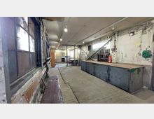 Ein einfacher, alter Werkstattraum mit Holzboden und abgenutzten Wänden. Im Raum befinden sich Metallwerkbänke, Regale und Leitungen an der Wand, mit einer Feuerlöscherstation an der rechten Seite.