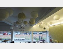 Raum mit großen Fenstern, die den Blick auf eine herbstliche Straße mit Autos und Laub ermöglichen. An der Decke hängen zahlreiche runde Papierlampen und ein moderner Kronleuchter; im Innenraum stehen Tische, Stühle und Pflanzen.