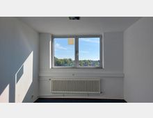 Ein leerer Raum mit weißen Wänden, einem großen rechteckigen Fenster in der Mitte, das Blick auf eine grüne Landschaft bietet. Unterhalb des Fensters befindet sich ein weißer Heizkörper, und der Boden ist mit dunkelblauem Teppich ausgelegt.