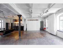 Industrie-inspirierter Raum mit weißen Wänden, einer glatten Decke und sichtbaren Balken. Links befindet sich eine kleine Bar mit Hockern, während am rechten Rand weiße Vorhänge und große Fenster erkennbar sind; der Boden ist abgenutzt und verleiht dem Raum ein rustikales Flair.