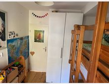 Kleines Kinderzimmer mit einem Etagenbett aus Holz rechts im Bild, einem weißen Kleiderschrank in der Mitte und einer Zimmertür mit einem Poster darauf. An der linken Wand hängen eine Weltkarte und ein Regal mit Spielsachen.
