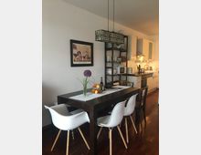 Essbereich in einer modernen Wohnung mit einem dunklen Holztisch, weißen Stühlen und einer Hängelampe aus schwarzem Draht. Im Hintergrund eine offene Küche mit weißen Schränken, einem Regal mit Geschirr und einer Arbeitsfläche mit verschiedenen Küchengeräten.