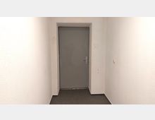 Ein schmaler, weiß gestrichener Flur mit einer grauen Tür am Ende. Der Boden ist mit dunkelgrauem Teppich ausgelegt, und an der rechten Wand befindet sich ein Lichtschalter.