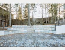 Gestufter Außenbereich mit Betonmauern und integriertem Beleuchtungssystem, zentraler Treppe und gepflastertem Boden, umgeben von hohen Bäumen. Im Hintergrund ist eine historische Steinmauer mit Rundbögen zu sehen.