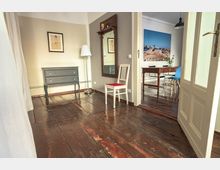 Holzfußboden-Flur mit hellbeigen Wänden, einer kleinen Kommode, einem Standspiegel und einem weißen Stuhl mit rotem Sitzpolster; durch eine offene Tür ist ein angrenzender Raum mit einem Holztisch, Stühlen und einem Landschaftsbild an der Wand sichtbar.