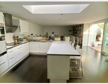 Moderne offene Küche mit weißen Schränken, dunklem Holzboden und einer großen Kücheninsel mit Barhockern. Im Hintergrund befindet sich ein Wohnbereich mit Bücherregalen, einer Treppe und einer großen Glasschiebetür, die zu einer Terrasse führt.