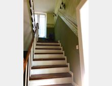 Treppenhaus in einem Innenraum mit Holzstufen und weißer Verkleidung, grün gestrichenen Wänden mit dekorativem Musterband und schwarzen Wandlampen. Oben links befindet sich ein Fenster mit weißen Vorhängen und ein Holzbettstuhl.