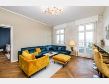 Modernes Wohnzimmer mit Fischgrätparkett, großem L-förmigem Sofa und gelbem Sessel samt Ottomane um einen Couchtisch. Hohe Fenster und eine Balkontür lassen viel Tageslicht herein; rechts steht eine schmale Theke mit Barhockern unter einer Deckenleuchte.