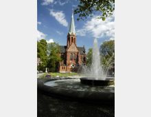 Backsteinkirche mit hohem, spitzem Turm und grüner Turmspitze, umgeben von Bäumen und einer gepflegten Parkanlage. Im Vordergrund ein großer Brunnen mit sprudelndem Wasser.
