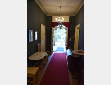 Schmaler Flur mit dunklen Wänden, einem roten Teppich und einem Kristall-Kronleuchter an der Decke. Der Raum führt zu einer Glastür mit roten Vorhängen, die den Blick auf einen Garten eröffnet; seitlich stehen Möbel wie Tische, Stühle und eine Bank.