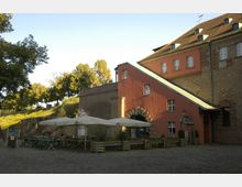 Außenansicht eines historischen Gebäudes mit rötlicher Fassade und Ziegeldach, umgeben von Bäumen. Im Vordergrund ist ein Biergarten mit Sonnenschirmen, Holztischen und Bänken auf einem gepflasterten Platz zu sehen.