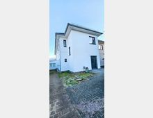 Zweistöckiges Wohnhaus mit weißer Fassade und dunkelgrauem Dach, seitlich ein gepflasterter Weg und eine kleine Grünfläche im Vordergrund. Das Gebäude hat mehrere Fenster und eine dunkelgraue Eingangstür.