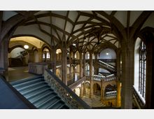 Historisches Innenraum eines Gebäudes mit gotisch inspirierten Merkmalen, einschließlich spitzbogiger Deckengewölbe, steinerner Geländer mit Ornamenten und Treppen, die verschiedene Ebenen verbinden. Große Bogenfenster lassen Tageslicht herein, und der Raum ist von braunen Sandsteinelementen geprägt.