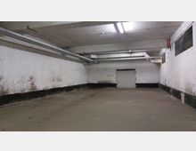 Leerer, karger Kellerraum mit Betonboden und weißen, teils verschmutzten Ziegelwänden. An der Decke verlaufen Rohre, und eine Ziegelwand mit zugemauerten Öffnungen ist im Hintergrund zu sehen.