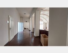 Ein heller Flur mit weißen Wänden und dunklem Boden, der zu einem offenen Bereich mit hohen, gewölbten Decken und großen Fenstern führt. Rechts ist ein Geländer mit einem Karton darauf zu sehen, links befinden sich geschlossene Türen und ein geöffneter Durchgang.