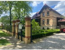 Historisches Haus mit der Aufschrift "Villa Luise 1877–2007" aus gelbem Backstein mit roten Verzierungen, umgeben von einem gepflegten Garten. Im Vordergrund ein schmiedeeiserner Zaun auf einer Mauer aus gelb-rotem Klinker und ein gepflasterter Weg mit Bäumen im Hintergrund.