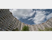 Hochhausanlage mit mehreren identischen Stockwerken in halbrunder Anordnung, mit Balkonen an jeder Wohnung und Baumkronen im Vordergrund unter einem teils bewölkten Himmel.