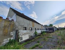 Ein verlassenes, einstöckiges Gebäude mit einem steilen Satteldach und verblasster Fassade, teils mit Holzplatten verschlossen. Im Vordergrund sind vereinzelt Pflanzen, eine herumliegende Tür und ein unbefestigter Weg sichtbar, während im Hintergrund ein weiteres kleines Gebäude steht.