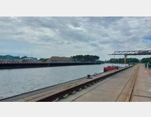 Hafenanlage mit einer breiten Betonpier und einem angrenzenden Hafenbecken, in dem ein rotes Frachtschiff liegt. Links sind große Holzstapel und industrielle Gebäude zu sehen, im Hintergrund stehen Bäume und technische Strukturen.