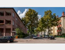 Wohnstraße mit mehrstöckigen Wohnhäusern auf beiden Seiten, teils in roten und gelben Farben. Bäume säumen die Straße, geparkte Autos stehen entlang des Gehwegs, und ein Fußgänger ist auf dem Bürgersteig zu sehen.