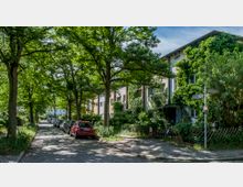 Baumbestandene Wohnstraße mit gepflastertem Gehweg und parkenden Autos am Straßenrand. Rechts stehen Reihenhäuser, von denen einige von dichtem Efeu bewachsen sind; ein Straßenschild zeigt "Waldhüterpfad".