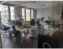 Ein modernes Büro mit mehreren weißen Schreibtischen, schwarzen Bürostühlen und Computerarbeitsplätzen. Große Fenster lassen Tageslicht herein und bieten Ausblick auf umliegende Gebäude; Zimmerpflanzen und Dekorationsgegenstände sind im Raum verteilt.