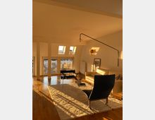 Helles Wohnzimmer mit großen Fenstern und Oberlichtern, durch die Sonnenlicht auf den Holzfußboden fällt. Der Raum ist möbliert mit einem cremefarbenen Sofa, einem schwarzen Sessel, einem Glastisch und einem hellen Teppich, ergänzt durch eine Wandleuchte und minimalistische Dekoration.