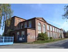 Backsteingebäude mit zwei Etagen, rechteckigen Fenstern mit Rolläden und einem Satteldach. Vor dem Gebäude befindet sich ein blaues Metalltor, umgeben von vereinzelten Pflanzen und einer gepflasterten Straße.