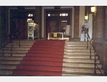 Treppenaufgang mit rotem Teppich, flankiert von Geländern, führt zu einem Eingang mit Glastüren und seitlich aufgestellten Schaukästen in einem historischen Gebäude. Wandlampen beleuchten den Bereich dezent.