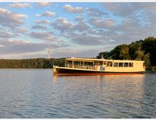 Ein weißes Ausflugsschiff schwimmt auf einem ruhigen See, umgeben von bewaldeten Ufern. Im Hintergrund ist ein großer Funkturm zu sehen, während der Himmel teils bewölkt ist und in warmem Licht erstrahlt.