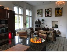 Geräumige Wohnküche mit einem Esstisch aus Holz und geflochtenen Stühlen, großen Fenstern mit Blick ins Grüne und einer stilvollen Kücheninsel mit Granitoberfläche. Retro-Werbeplakate hängen an der weißen Wand, und ein Kronleuchter hängt von der Decke.