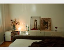 Ecke eines Wohnzimmers mit einer weißen Kommode, darauf eine Vase mit Trockenblumen, eine kleine Lampe, zwei gerahmte Bilder und Dekorationsobjekte. Im Vordergrund ist ein Bett mit einer braunen Decke und hellem Bettbezug zu erkennen.