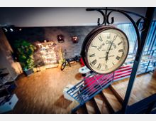 Innenraum im Industrial-Stil mit freitragender Metalltreppe und Holzfußboden; im Vordergrund hängt eine große runde Bahnhofs-Wanduhr. Im Hintergrund sind eine Backsteinwand, dekorative Beleuchtung und ein Fahrrad als Einrichtungselement zu sehen.