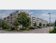 Wohnblock in einer Wohnsiedlung mit mehreren Stockwerken und Balkonen, umgeben von Bäumen und parkenden Autos. Eine asphaltierte Straße mit Fußwegen und Straßenlaternen führt am Gebäude vorbei.