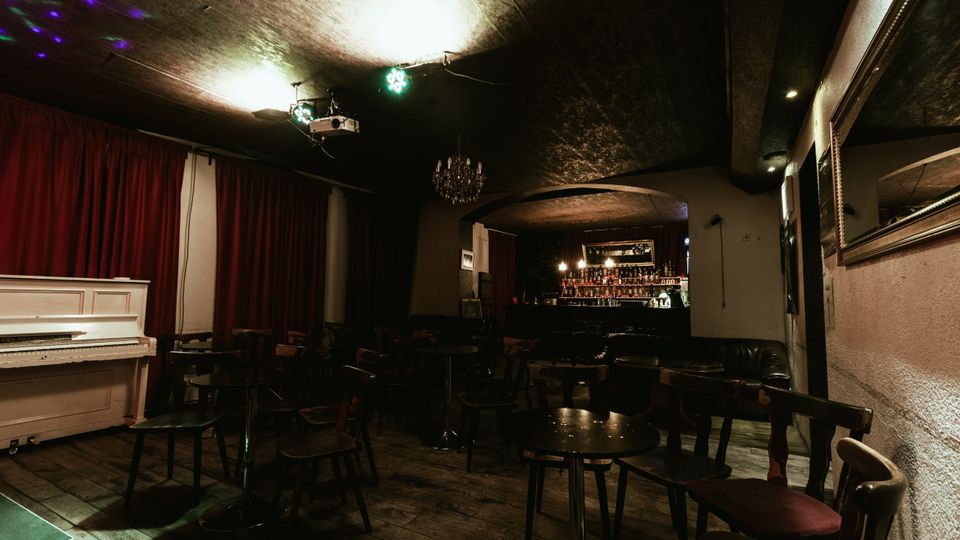 Dimly lit lounge or small music venue interior with round tables and wooden chairs on a dark wood floor, a white upright piano by red curtains, and a bar with bottles under an arched opening. Low ceiling lighting and a chandelier add to the intimate, basement-like setting.