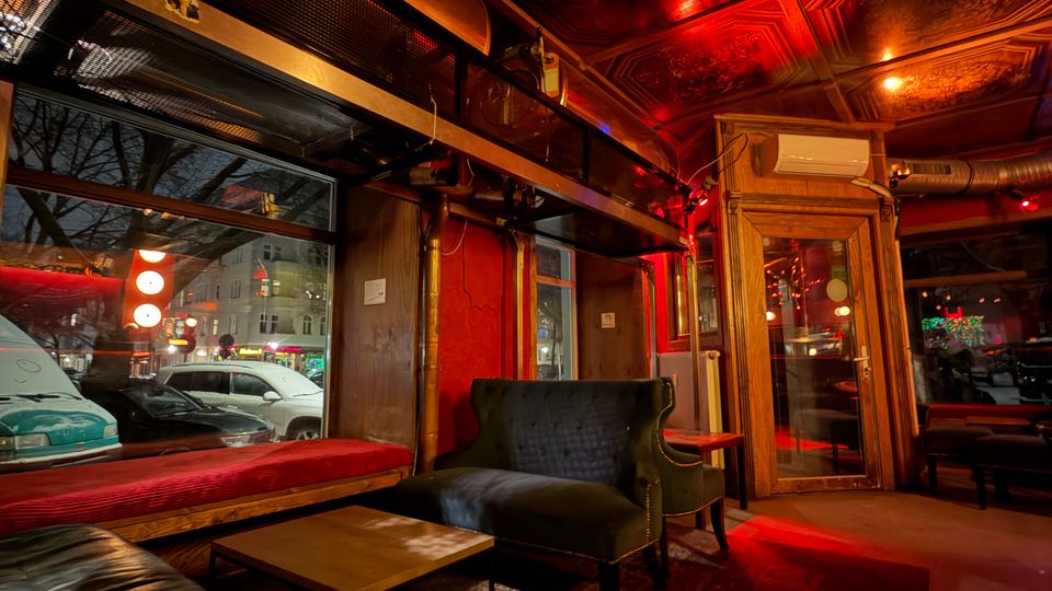 Innenraum einer Bar-Lounge mit dunklen Ledersofas und Sesseln um niedrige Tische auf einem gemusterten Teppich, neben großen Schaufenstern zur Straße. Kassettierte Decke mit ornamentalen Feldern und warmem, rötlichem Licht sowie Holzrahmen und Glastür im Hintergrund.