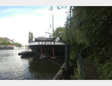 Ein festgemachtes Schiff mit dem Namen "Kurier" liegt an einem Flussufer, umgeben von Bäumen und dichtem Grün. Im Hintergrund sind Gebäude, ein weiterer kleineres Boot und eine Brücke über den Fluss zu sehen.