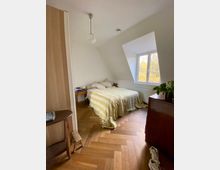 Ein Schlafzimmer mit einem niedrigen Bett, das mit einer gelb-weiß gestreiften Decke bedeckt ist, steht unter einer Dachschräge mit einem großen Fenster. Der Raum hat einen Fischgrät-Holzboden, eine weiße Hängeleuchte und Möbel wie einen Nachttisch und eine Kommode.