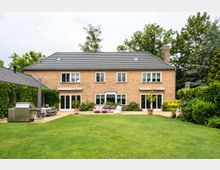 Zweistöckiges Einfamilienhaus aus rotbraunem Backstein mit grauem Satteldach, großen Fenstern und Terrassentüren, umgeben von einem gepflegten Garten mit Rasen, Terrassenmöbeln und grüner Bepflanzung. Im Vordergrund steht eine Grillecke und ein Sitzbereich, im Hintergrund sind hohe Bäume sichtbar.