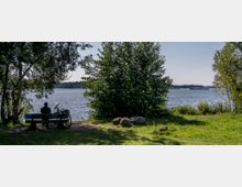 Ein Mann sitzt auf einer Parkbank am Ufer eines Sees, neben ihm steht ein Fahrrad. Im Vordergrund ist eine grasbewachsene Fläche mit Felsen, ringsum von Bäumen und Sträuchern umgeben, während im Hintergrund der See und ein frachtähnliches Boot zu sehen sind.