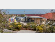 Modernes Veranstaltungsgebäude mit Flachdach und einer Fassade in Beige mit großen Glasfronten, umgeben von grünem Buschwerk und Gehwegen. Im Hintergrund ist eine Kleinstadt mit Wohnhäusern sowie ein hoher Funkmast sichtbar.