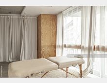 Behandlungsraum mit einem beige gepolsterten Massagetisch auf Holzbeinen, umgeben von halbtransparenten Vorhängen und großer Fensterfront, die Tageslicht hereinlässt. Im Hintergrund stehen ein Wandschrank aus Holz und weitere bodenlange Vorhänge.