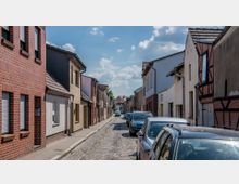 Schmale Kopfsteinpflasterstraße in einem Wohngebiet mit zweistöckigen Häusern aus Backstein und Putzfassaden, geparkt sind Autos entlang beider Straßenseiten. Der Himmel ist teilweise bewölkt.