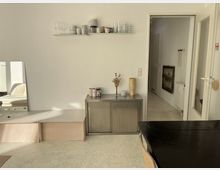 Minimalistisch eingerichteter Innenraum mit weißen Wänden und hellem Boden. Ein Regal mit Gläsern und Tassen hängt an der Wand, darunter steht ein Metallschrank mit Deko-Objekten. Rechts führt eine offene Tür in einen Flur, links ein Spiegel, der ein Sofa reflektiert.