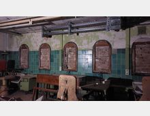Ein verlassenes Zimmer mit abblätternder grüner Farbe an den Wänden, grün gefliesten unteren Wandbereichen und zugemauerten, bogenförmigen Fenstern. Im Raum befinden sich alte Möbel und Geräte, darunter ein Stuhl, Tische und Schränke.