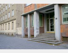 Eingang eines Wohn- oder Bürogebäudes mit einer kleinen Treppe, umgeben von einer Fassade aus hellbeigen und orangefarbenen Fliesen. Die Glasfenster und die Tür sind von Betonpfeilern eingerahmt, teils mit Graffiti bedeckt.