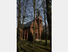 Backsteinkirche mit roter Fassade und einem hohen spitzen Turm, umgeben von Bäumen und einer Rasenfläche mit Bänken im Vordergrund.
