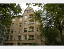 Wohngebäude im Stil der Gründerzeit mit cremefarbener Fassade, mehreren Balkonen und symmetrisch angeordneten Fenstern, umgeben von Bäumen in einer grünen, parkähnlichen Umgebung.