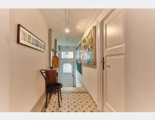 Schmaler, heller Flur in einem Wohngebäude mit hellen Wänden, einer weißen Kassettendecke und einem gemusterten Fliesenboden. An den Wänden hängen Bilder, und im Hintergrund führt eine Treppe zu einer verzierten Glastür mit Oberlicht.